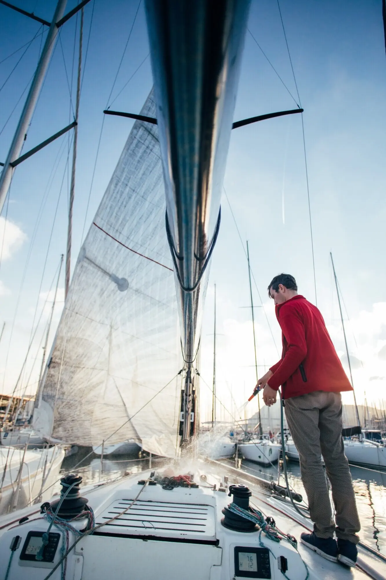 A sailboat owner or yachtsman uses a hose to rinse salt water off the yacht’s deck while docked or parked in a marina at sunset.